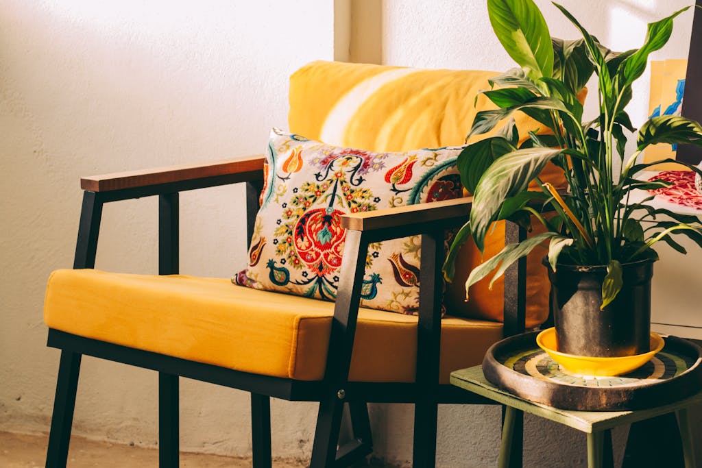 A bright, inviting corner with a yellow armchair, colorful pillow, and lush green plant in sunlight.