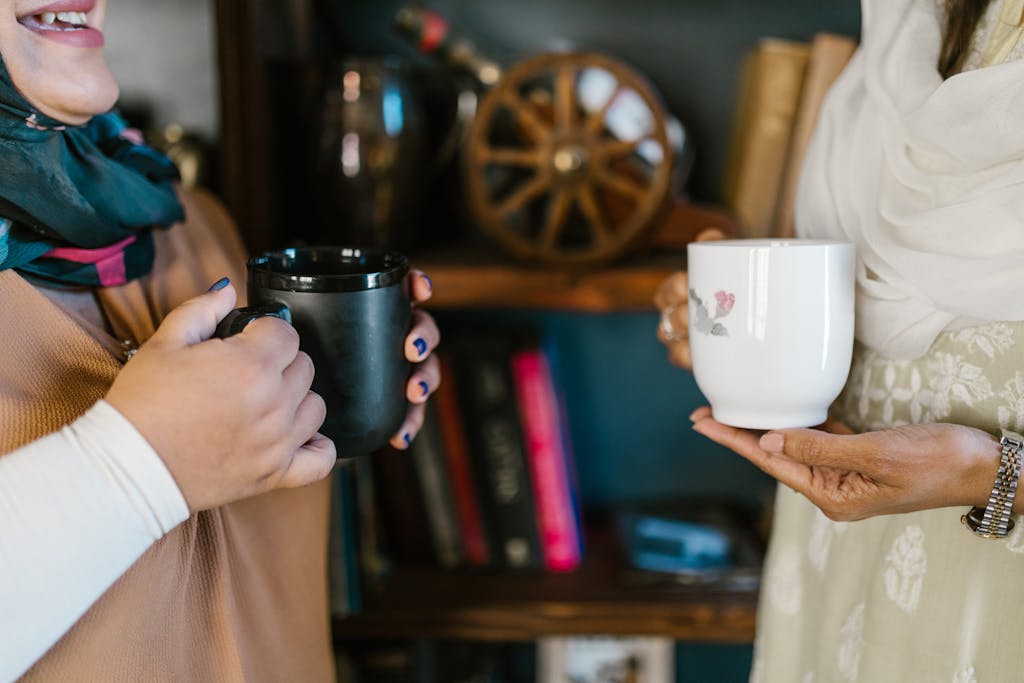 Two women holding mugs share a warm moment indoors. Close-up of hands and cups.